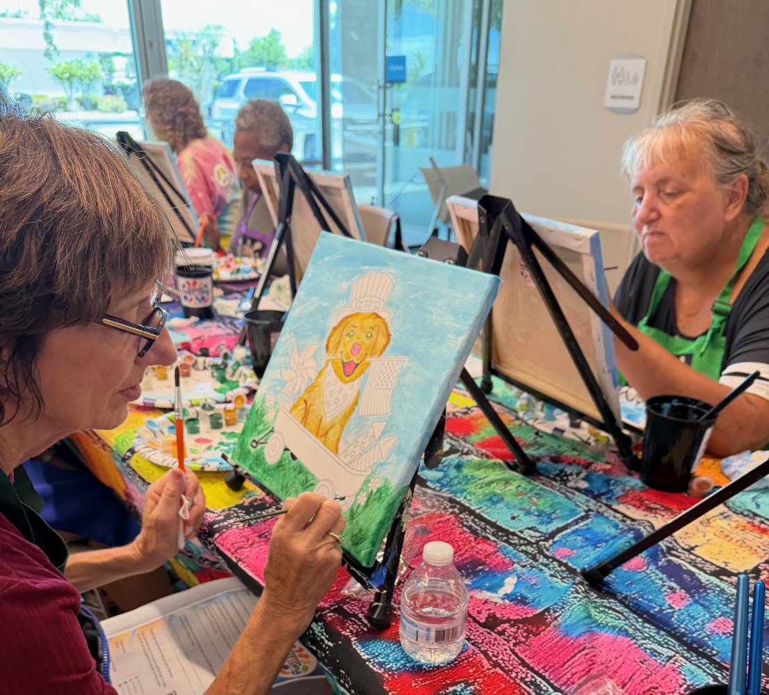 Older women participating in a painting class, each working on colorful canvases, with one woman painting a cheerful dog in a bathtub.