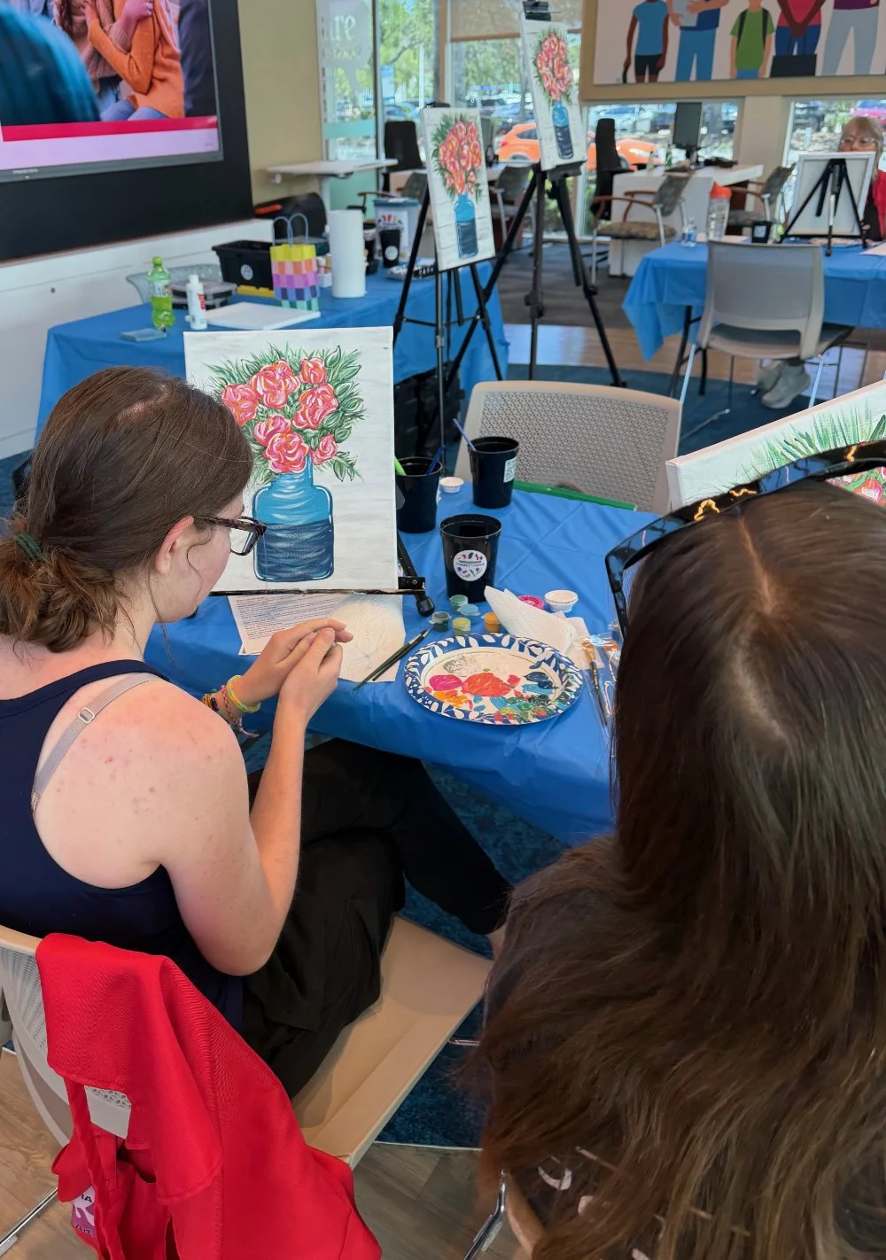 People participating in a painting class creating paintings of pink flowers in a blue vase, with art supplies on a blue-covered table in a bright room with large windows.