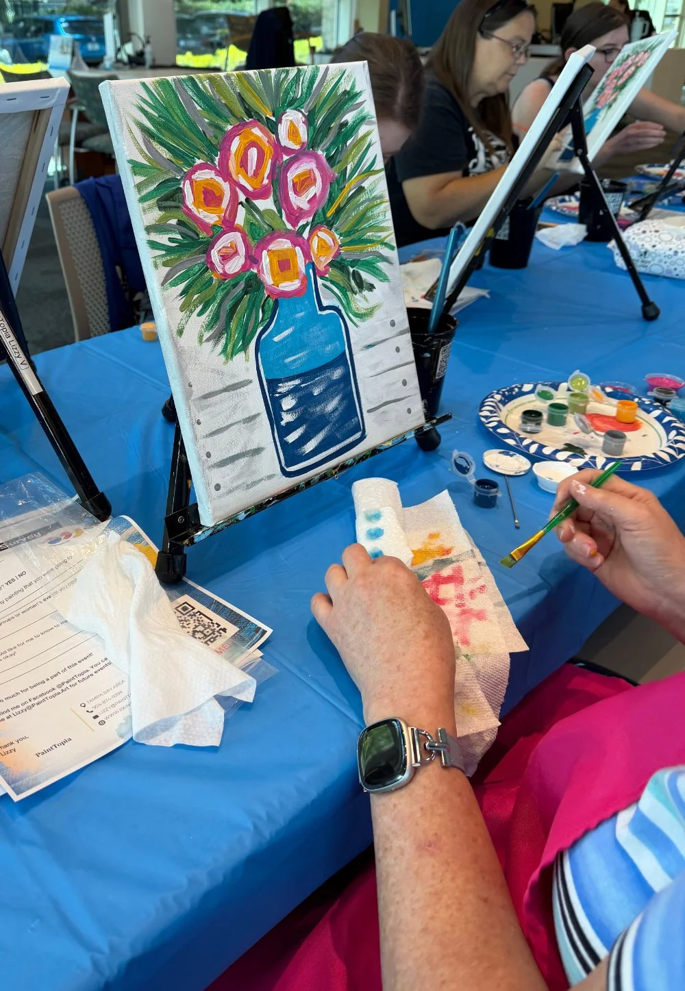 Painting of pink and orange flowers in a blue vase on a canvas with green leaves, set on an easel during a group painting activity.