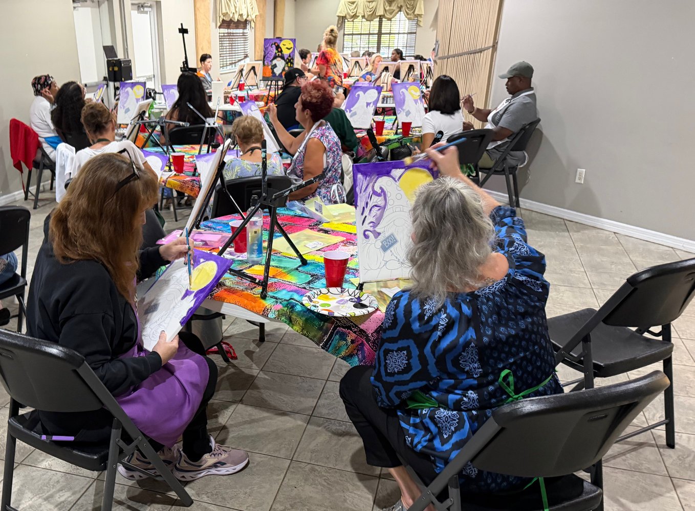 A group of people participating in a painting class around tables covered with a rainbow-colored tablecloth. They are painting canvases featuring a white character with a moon and purple background. The room has beige tiled floors, grey walls, and la