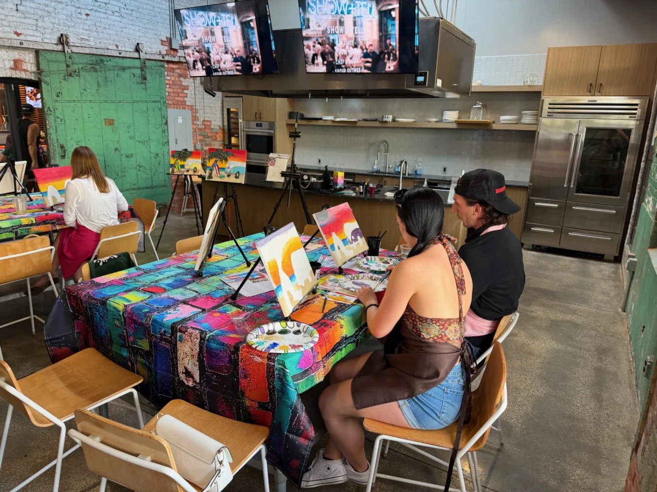 People participating in a canvas painting class in a modern, industrial-style room with colorful tablecloths, paintings of tropical scenes, and multiple canvas paintings on display.