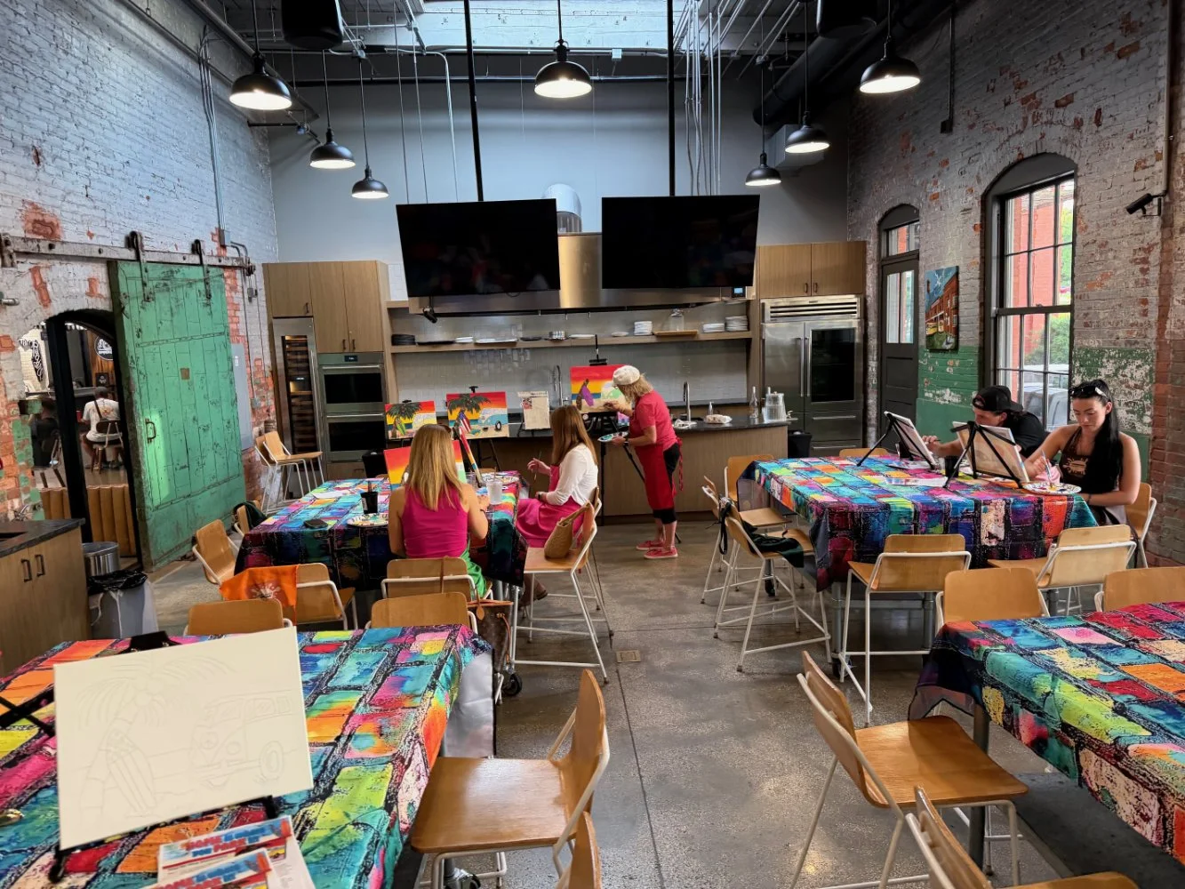 People sitting at tables in a colorful art studio or cafe, painting on canvases, with a kitchen area in the background and large windows on the right side.