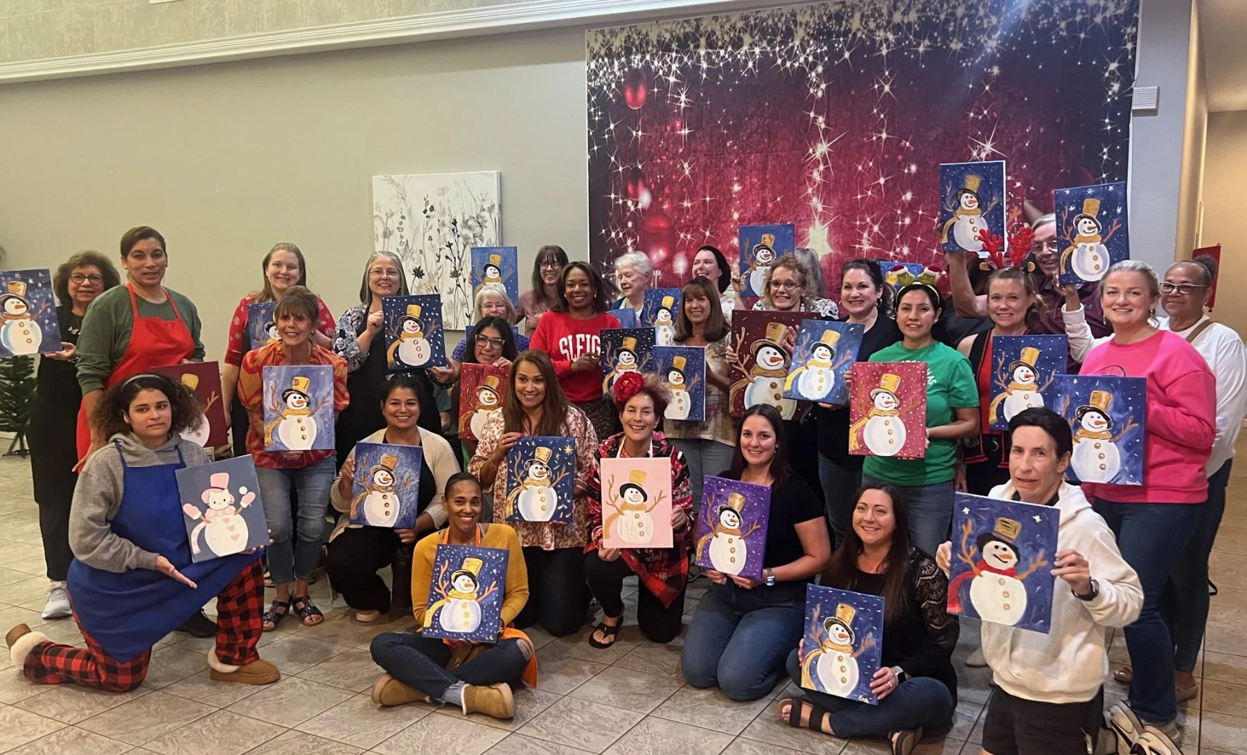 A group of women and a few men holding up decorated holiday artwork featuring snowmen, in a room with a festive backdrop and Christmas-themed decorations.