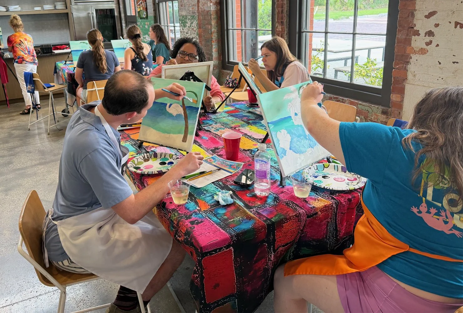 People participating in a group painting activity in an indoor space with large windows and exposed brick walls, sitting at a table covered with a colorful tablecloth, each working on a canvas.
