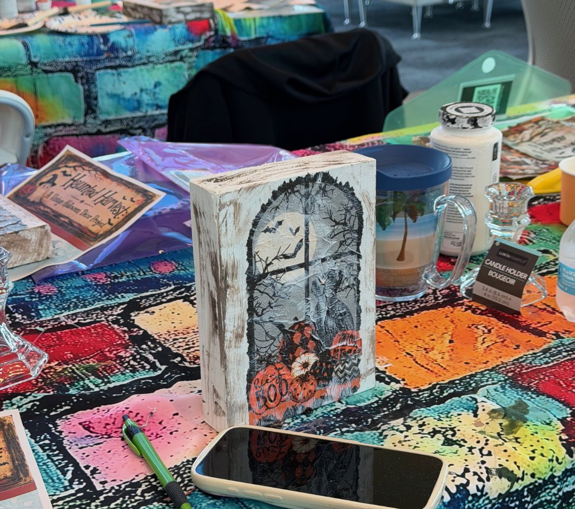 Decorative Halloween-themed wood sign with a silhouette of a haunted house, bats, and pumpkins on a multicolored tablecloth at a craft or market display.
