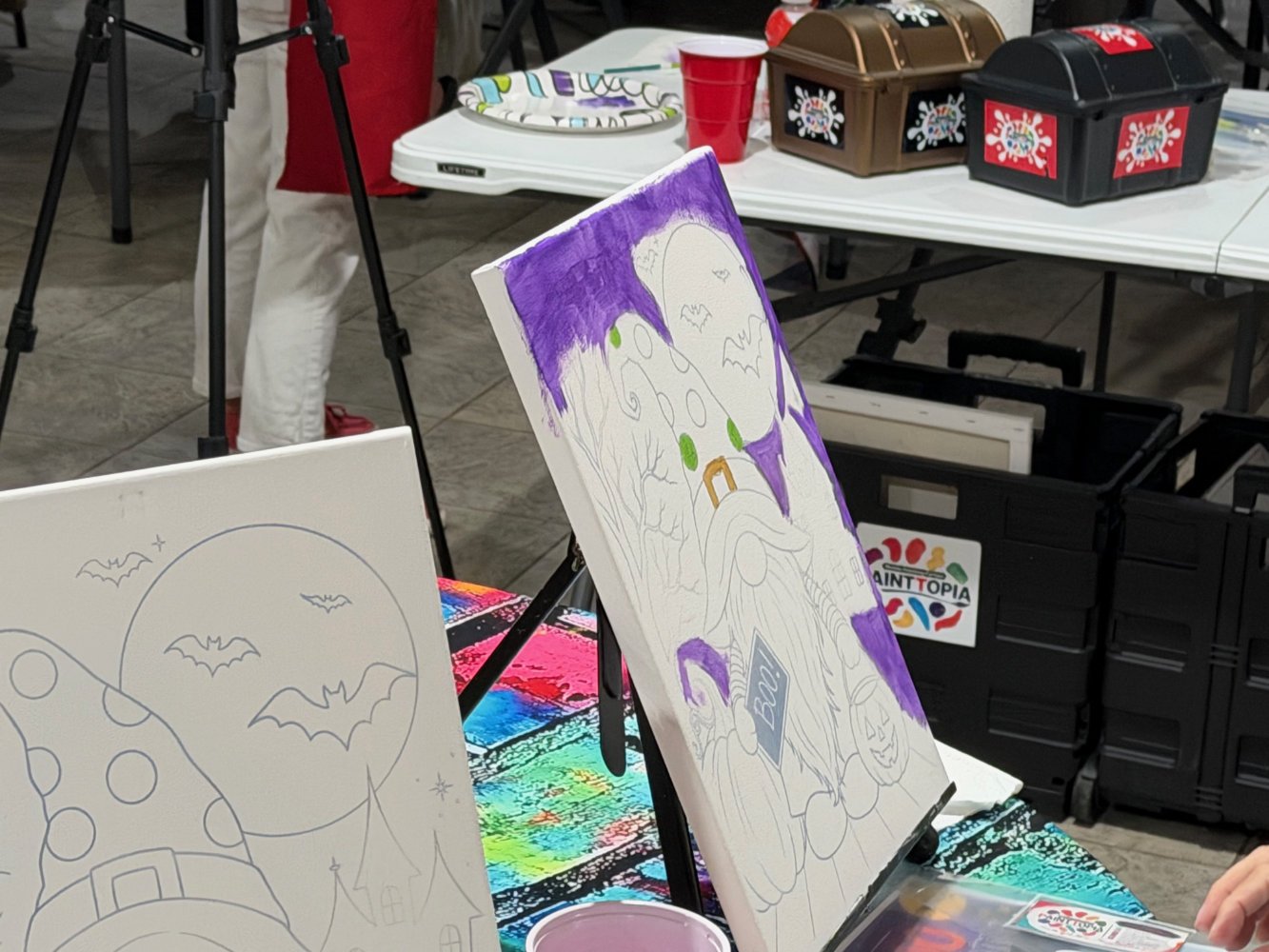 Unfinished Halloween-themed paintings on canvases, a table with art supplies, and storage bins at an art event or craft fair.