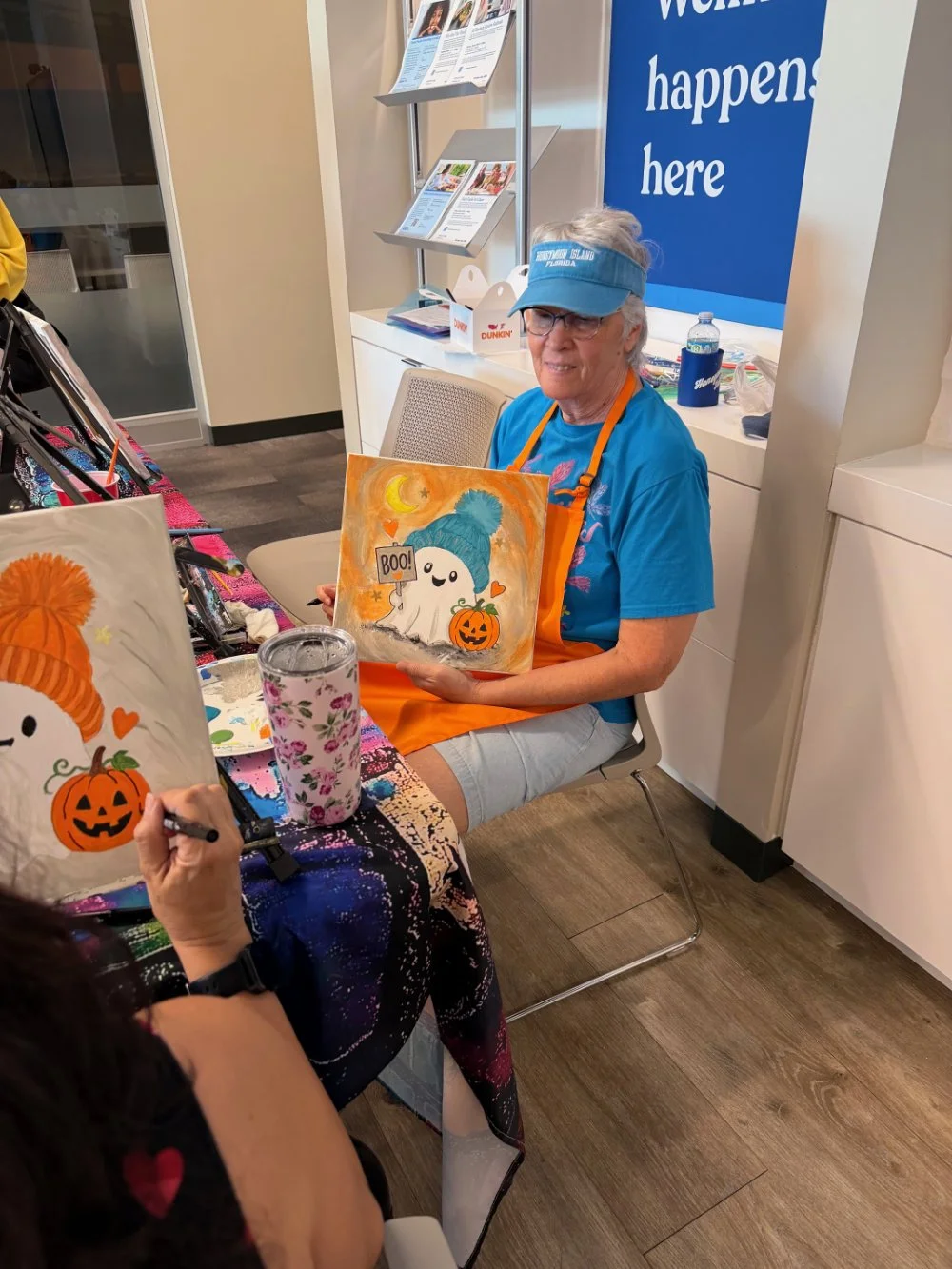 A woman wearing glasses, a blue visor, and an orange apron is sitting at a table displaying Halloween-themed artwork. She is holding a painting of a ghost with a blue hat, a small pumpkin, and a 'BOO!' sign. There are other paintings on the table, an