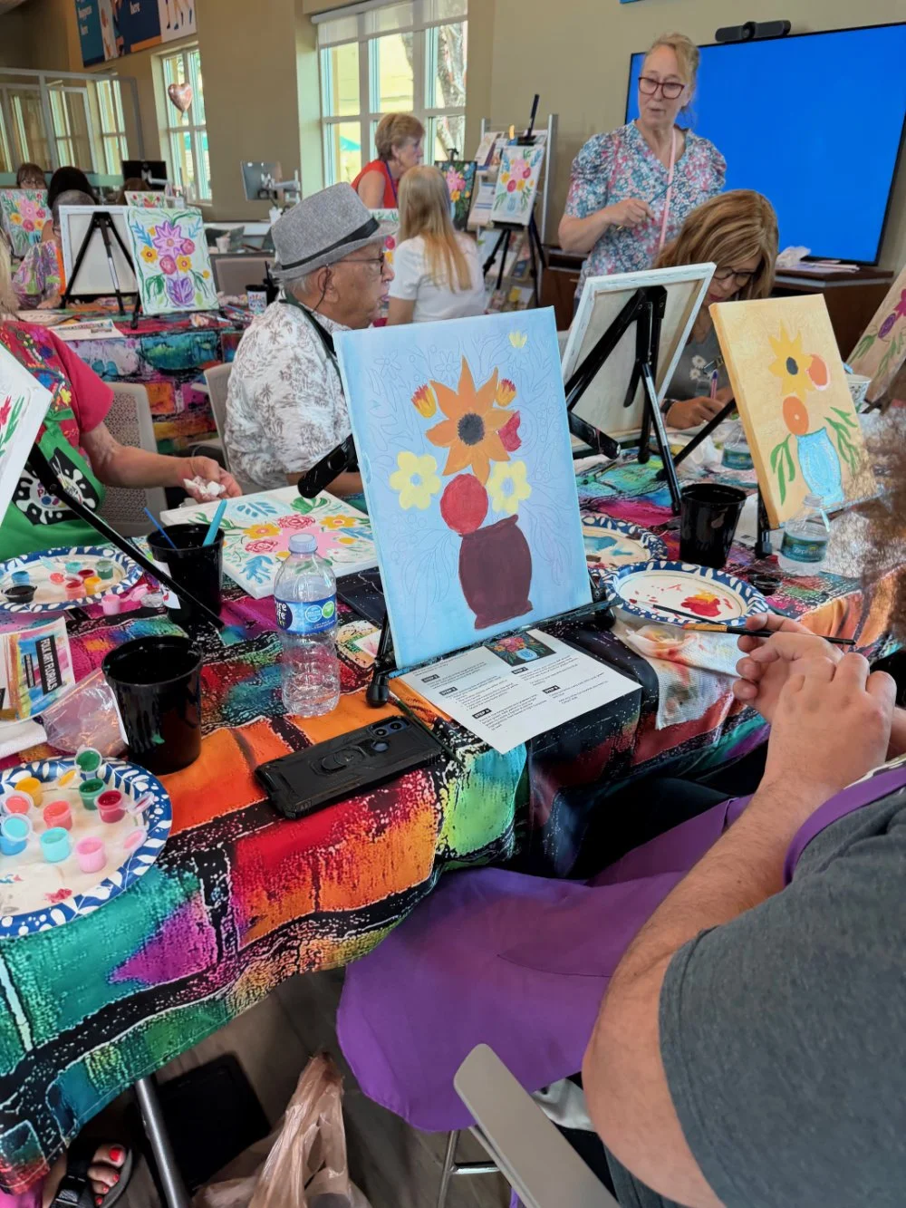 A group of people participating in a painting class, creating colorful flower paintings on canvases set on easels. The room is bright with large windows, and the table is covered with a vibrant, multicolored cloth.