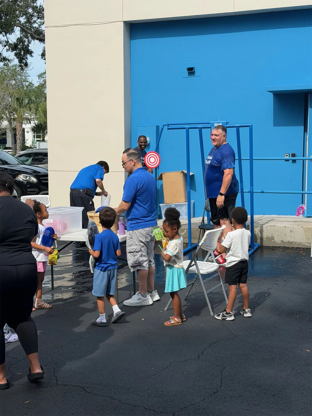 Children and adults lining up for a dunk tank game outside a building, with a man in the dunk tank smiling, and another man preparing items at a table.