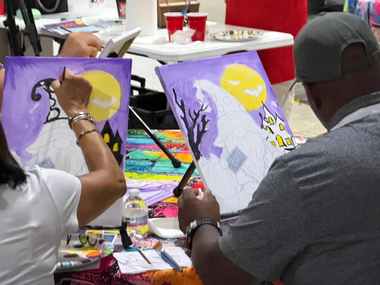 People painting Halloween-themed pictures on canvases with a moon, haunted house, and bats, using colorful paints at a table with art supplies.