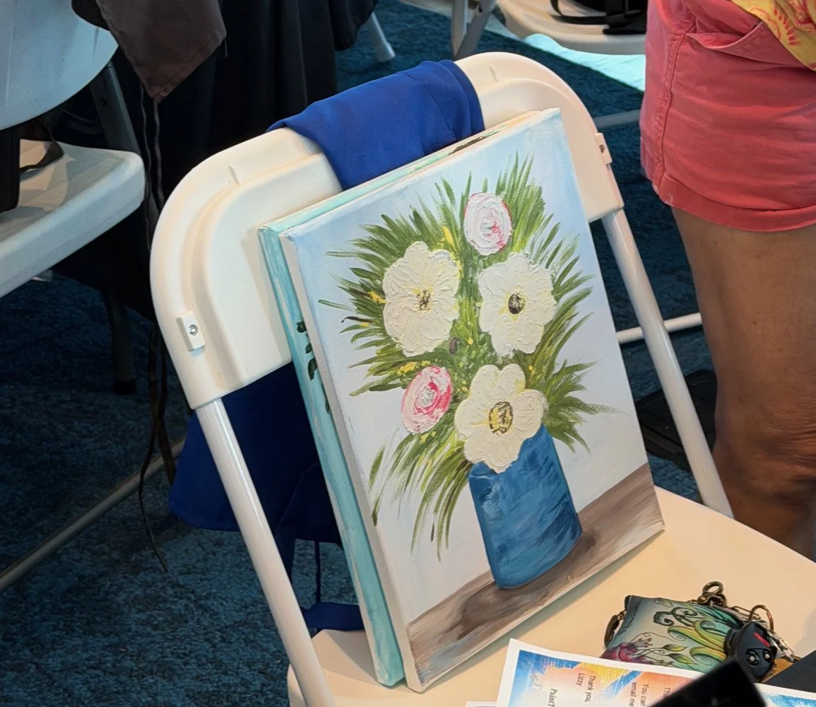 Painting of white and pink flowers in a blue vase on a table, propped against a white chair, with a person standing nearby.