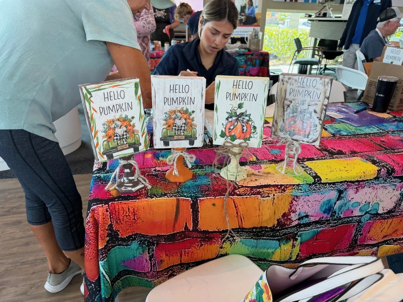 A display table at a craft fair with decorative signs featuring fall pumpkin themes, colorful tablecloth, and people browsing in the background.