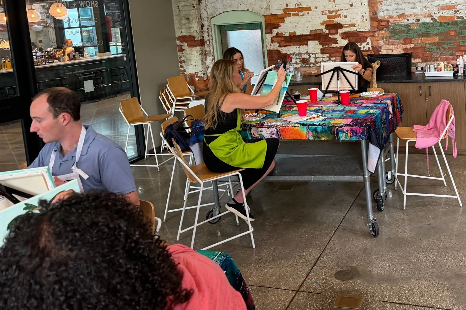 Four women sitting at a table in a cafe, one of them painting on a canvas, with a colorful tablecloth and red cups, exposed brick wall and a glass door in the background.