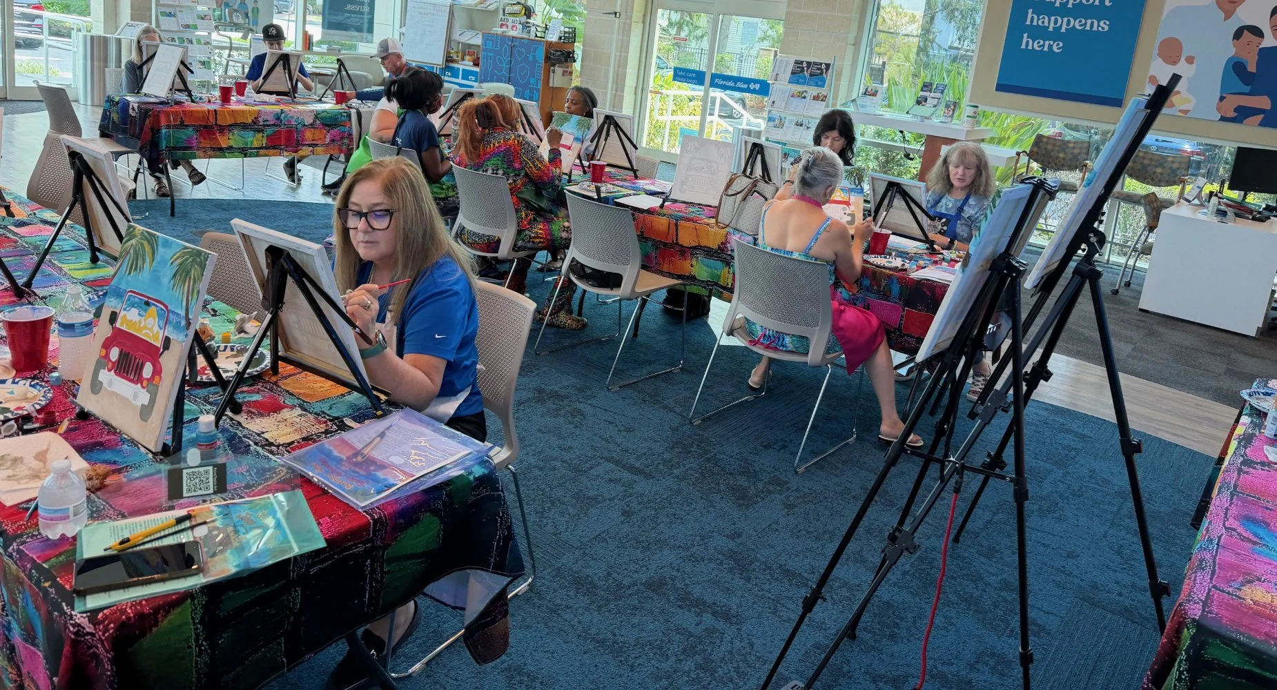People participating in a painting class at tables covered with colorful tablecloths, with canvases displaying various painted scenes, inside a bright room with large windows and art supplies.