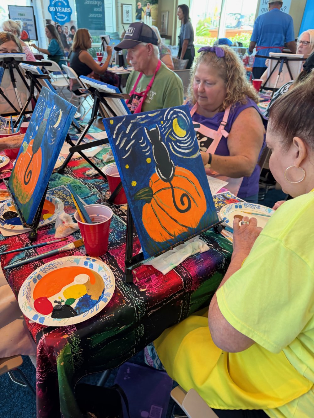 People participating in a painting class, working on canvases with colorful, Halloween-themed designs, such as pumpkins and black cats, at a table with paint palettes and brushes.