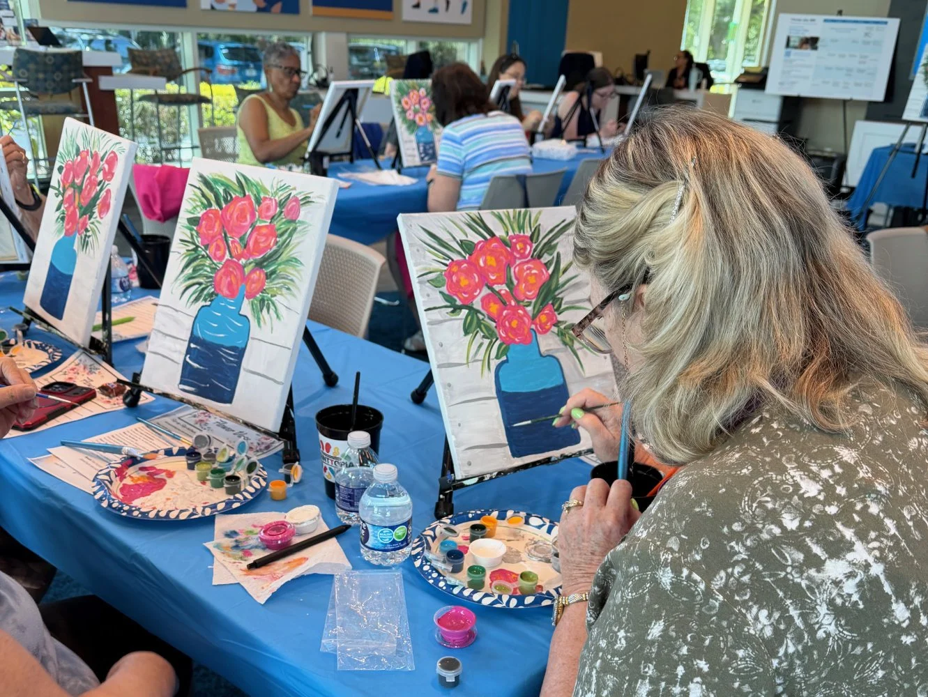 A group of people participating in a painting class, creating flower arrangements in blue vases on canvases, with art supplies and water bottles on the table.