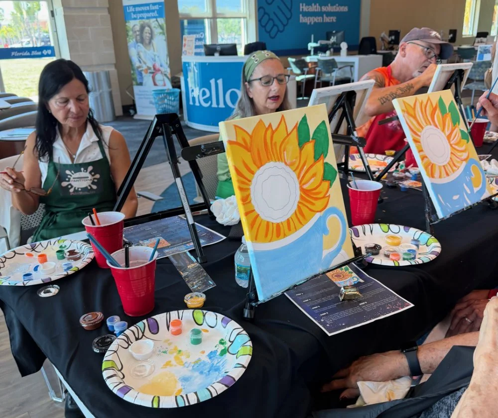 People participating in a paint and sip art class, painting sunflower-themed paintings on canvases, with painting supplies and drinks on the table.