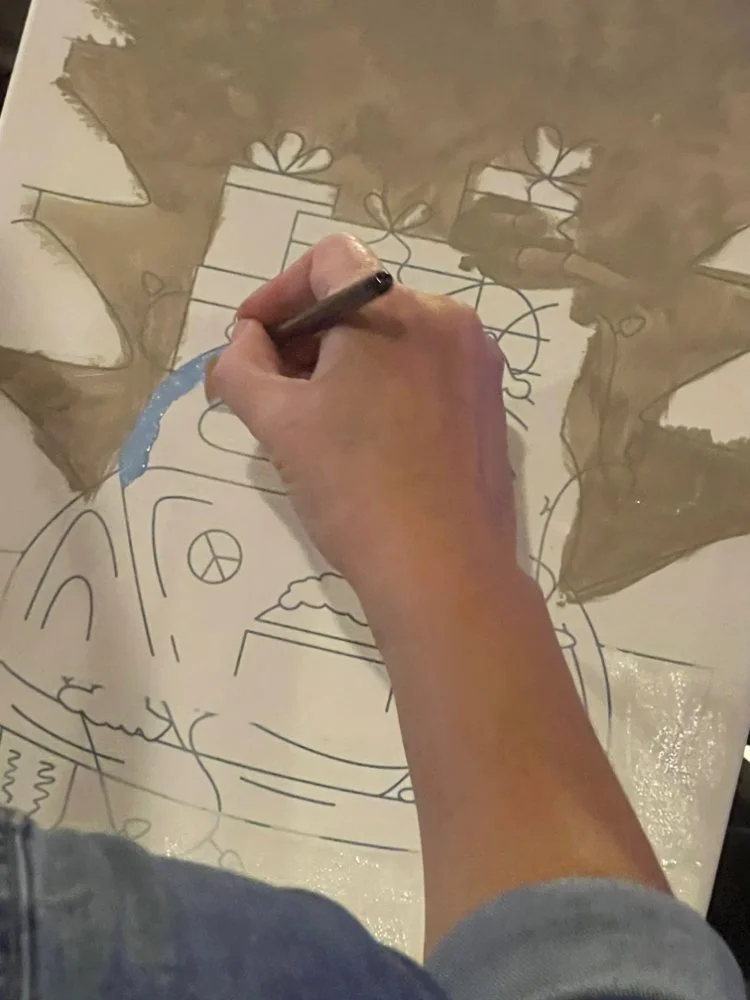 A person is drawing a festive holiday scene with presents, Christmas trees, and other holiday decorations on a large sheet of paper.