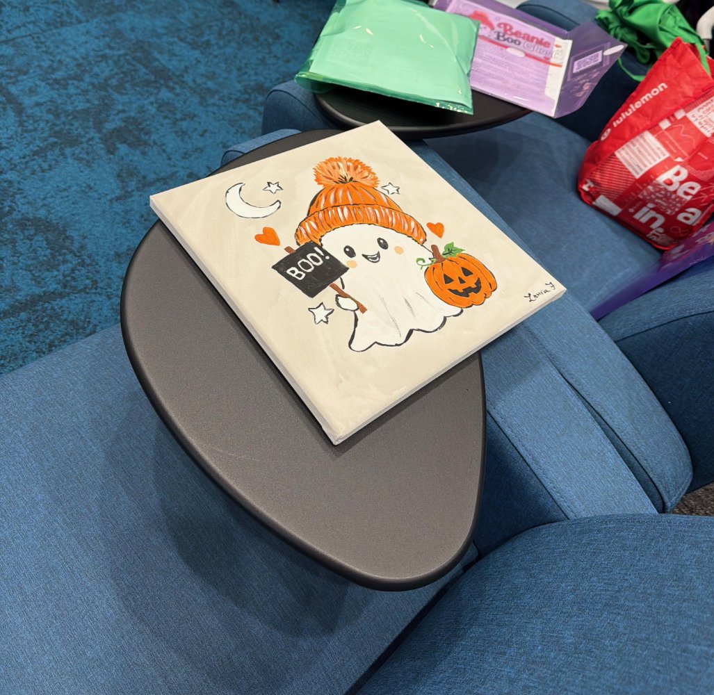 A greeting card with a cheerful cartoon ghost wearing an orange pumpkin hat, holding a small pumpkin bucket, surrounded by moon and star illustrations, on a dark gray oval table in a blue upholstered seating area with various colorful items in the ba