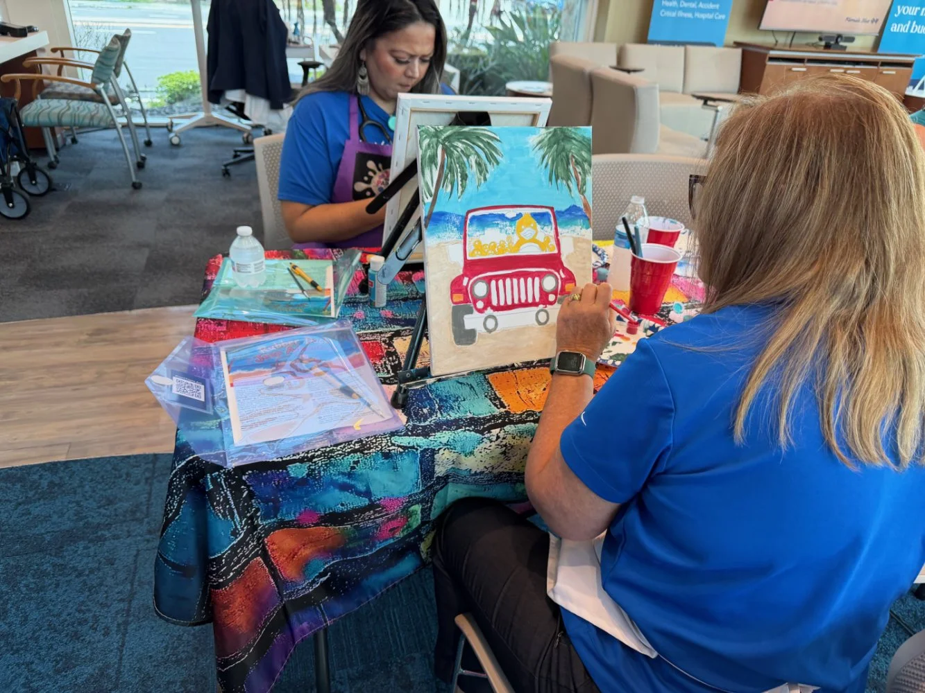 Two women painting tropical beach scenes on canvas at a table in a bright indoor setting. One woman, with blonde hair, is in focus with her back to the camera, wearing a blue shirt, while the other woman, with darker hair, works on her art in the bac