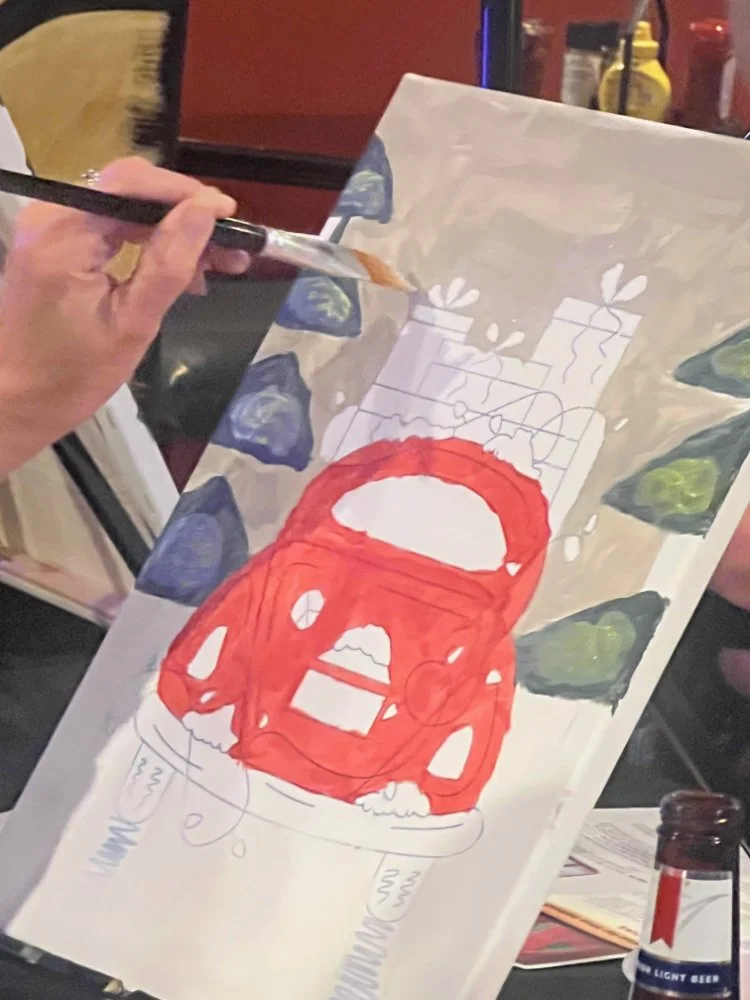 Person painting a colorful illustration of a red fire truck with trees, presents, and snowflakes on a canvas in a restaurant setting.