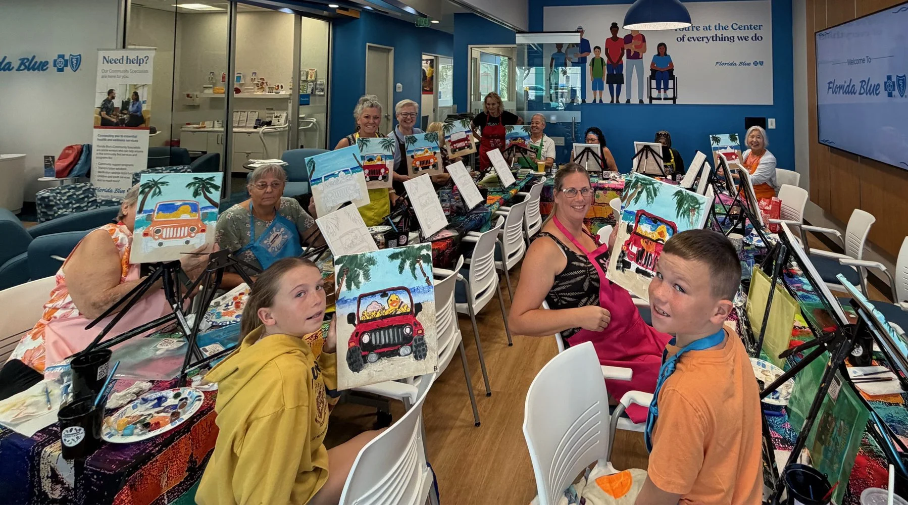A group of children and adults participating in a painting activity at tables with art supplies, showcasing colorful beach and vehicle-themed paintings, inside a community center with bright decor.