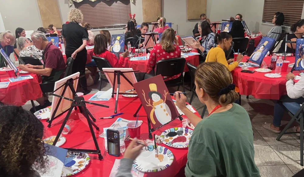 Group of people painting Christmas-themed snowmen on canvases at a holiday painting event in a room with red tablecloths.