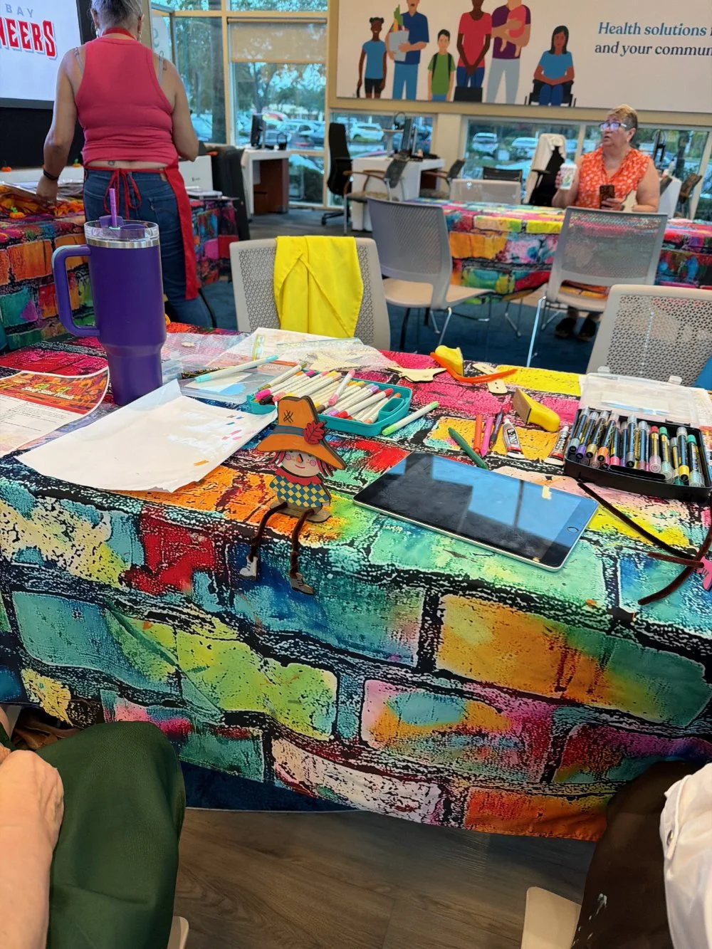 A colorful table with art supplies, markers, pens, a tablet, and a paper sketch in a brightly lit community room with several people in the background.