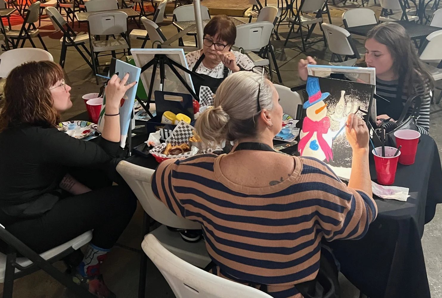 Four women sitting around a table at a party, painting holiday-themed decorations, with food and drinks on the table.