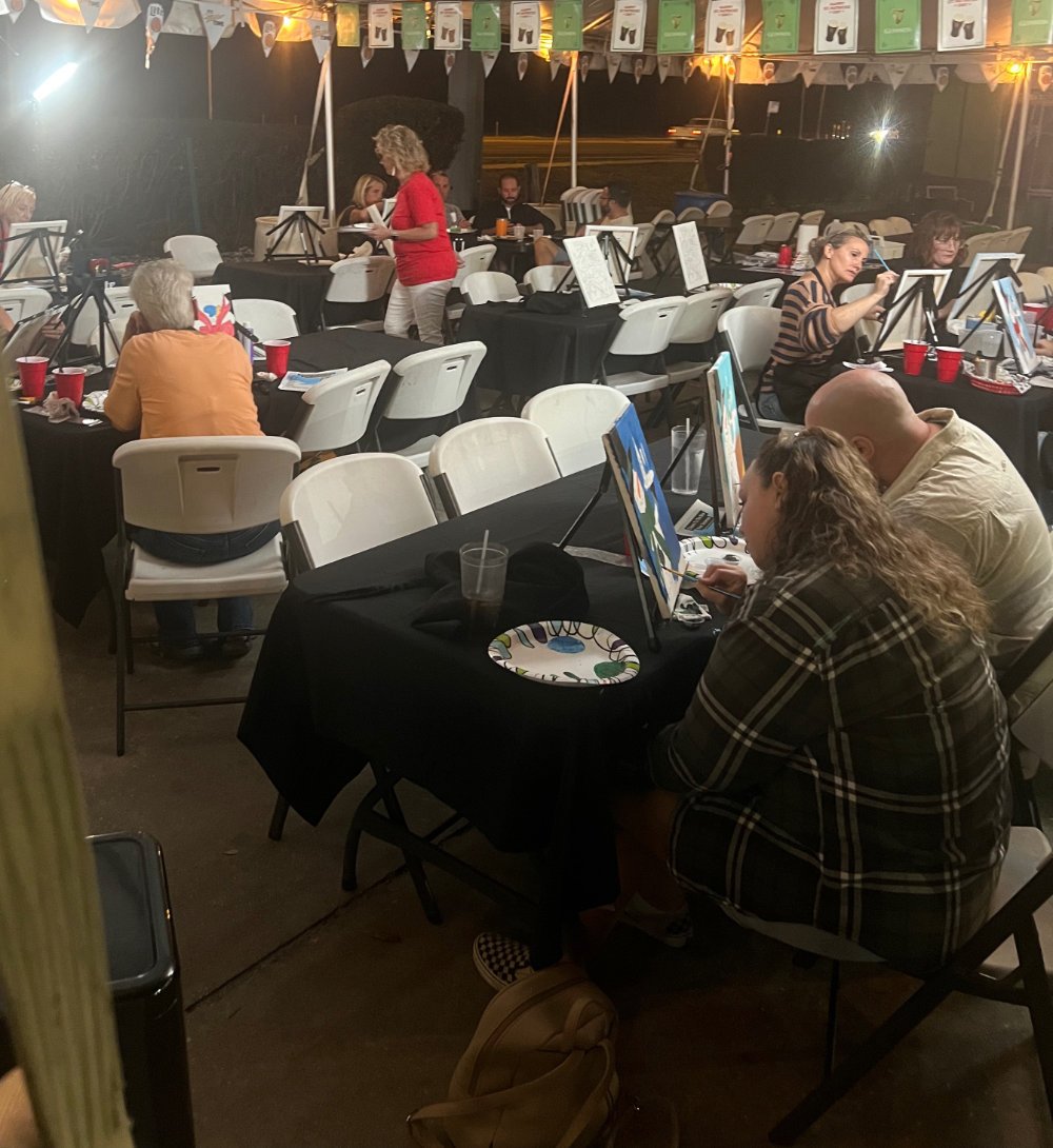 People participating in a painting class at night, seated at tables with easels and painting supplies, under string lights and colorful banners.