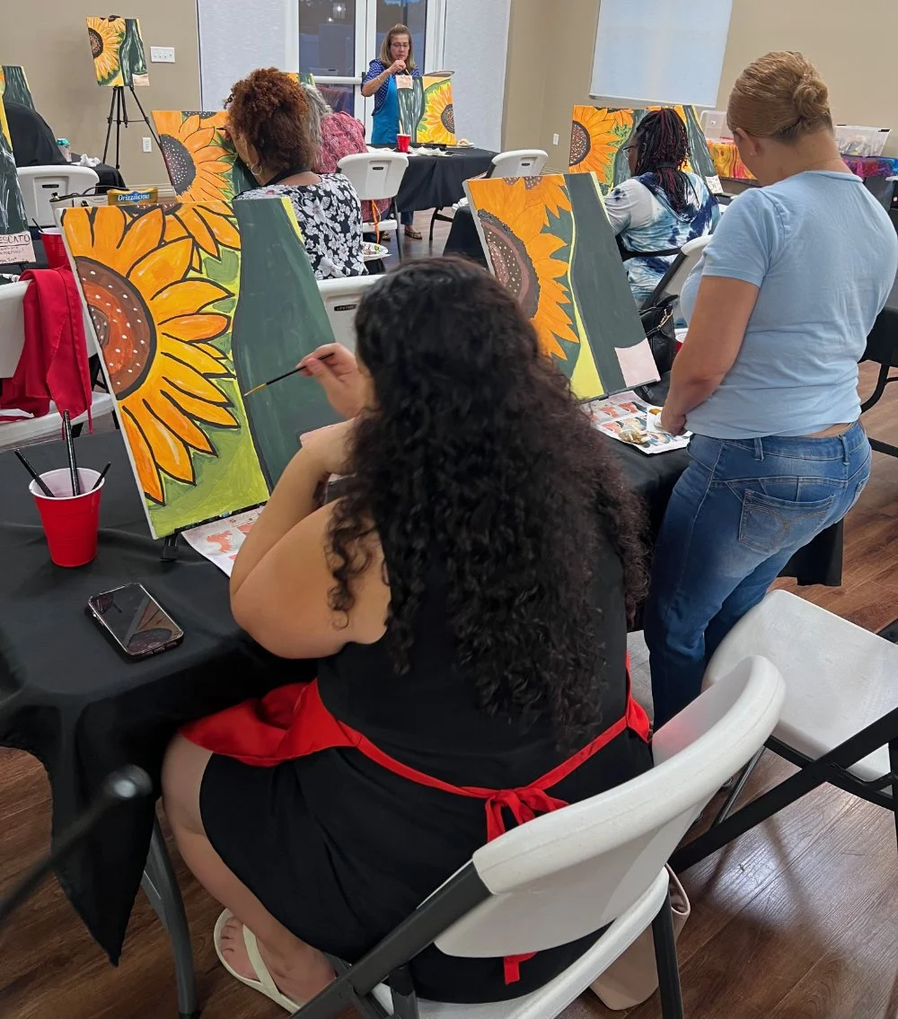 People participating in a painting class creating sunflower artwork. Some individuals are painting while others observe. A woman in the foreground is painting a sunflower on a canvas, and a woman standing nearby is watching.