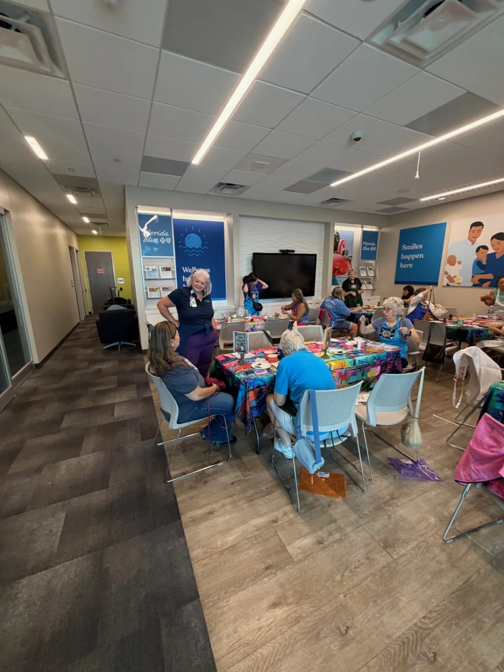 Group of elderly women and staff members gathered in a community room decorated with bright, colorful tablecloths, engaged in an arts and crafts activity.
