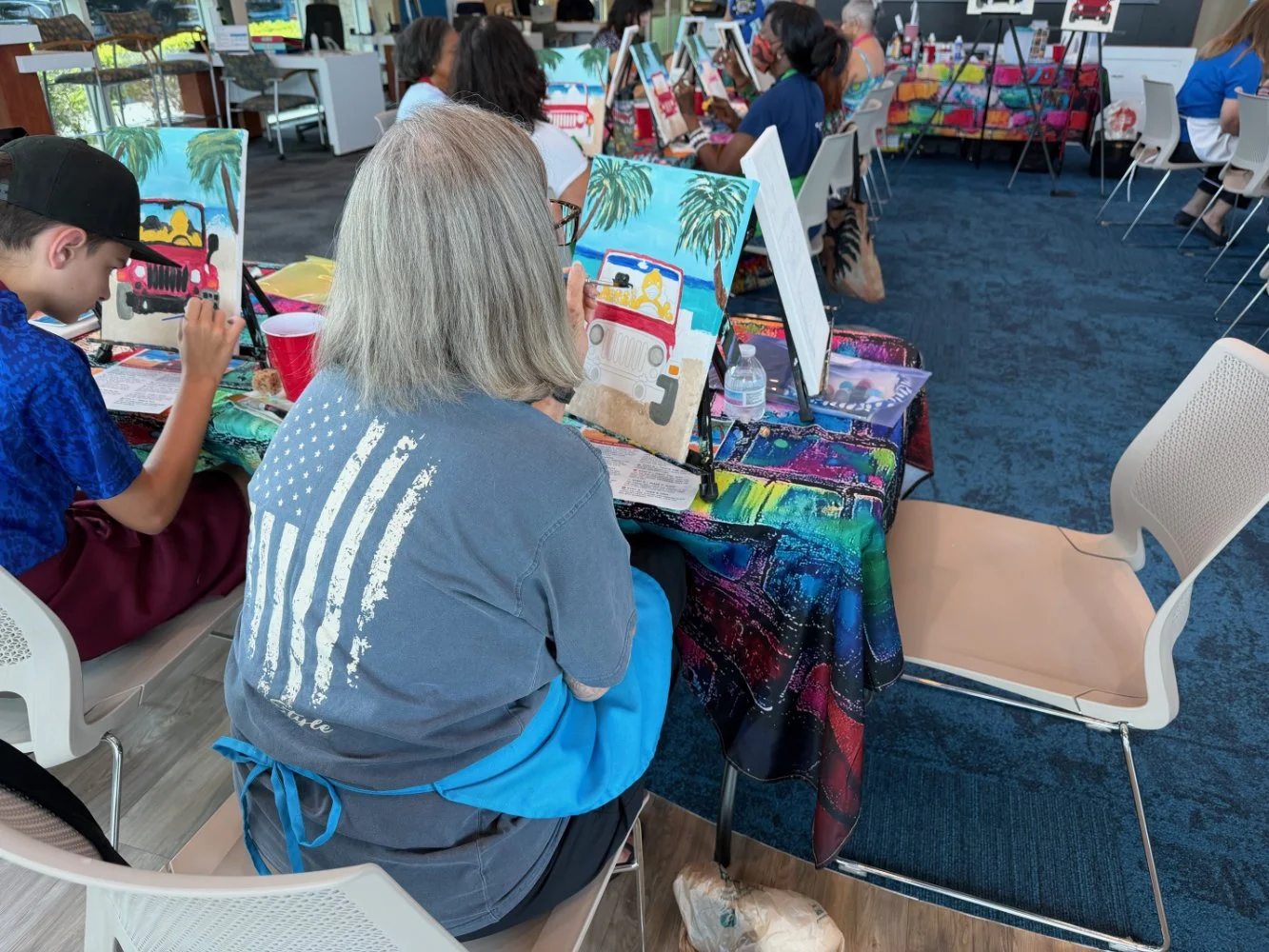 People participating in a painting class, working on canvases depicting beach scenes with palm trees, ocean, and vehicles, seated at colorful tables in an indoor setting.