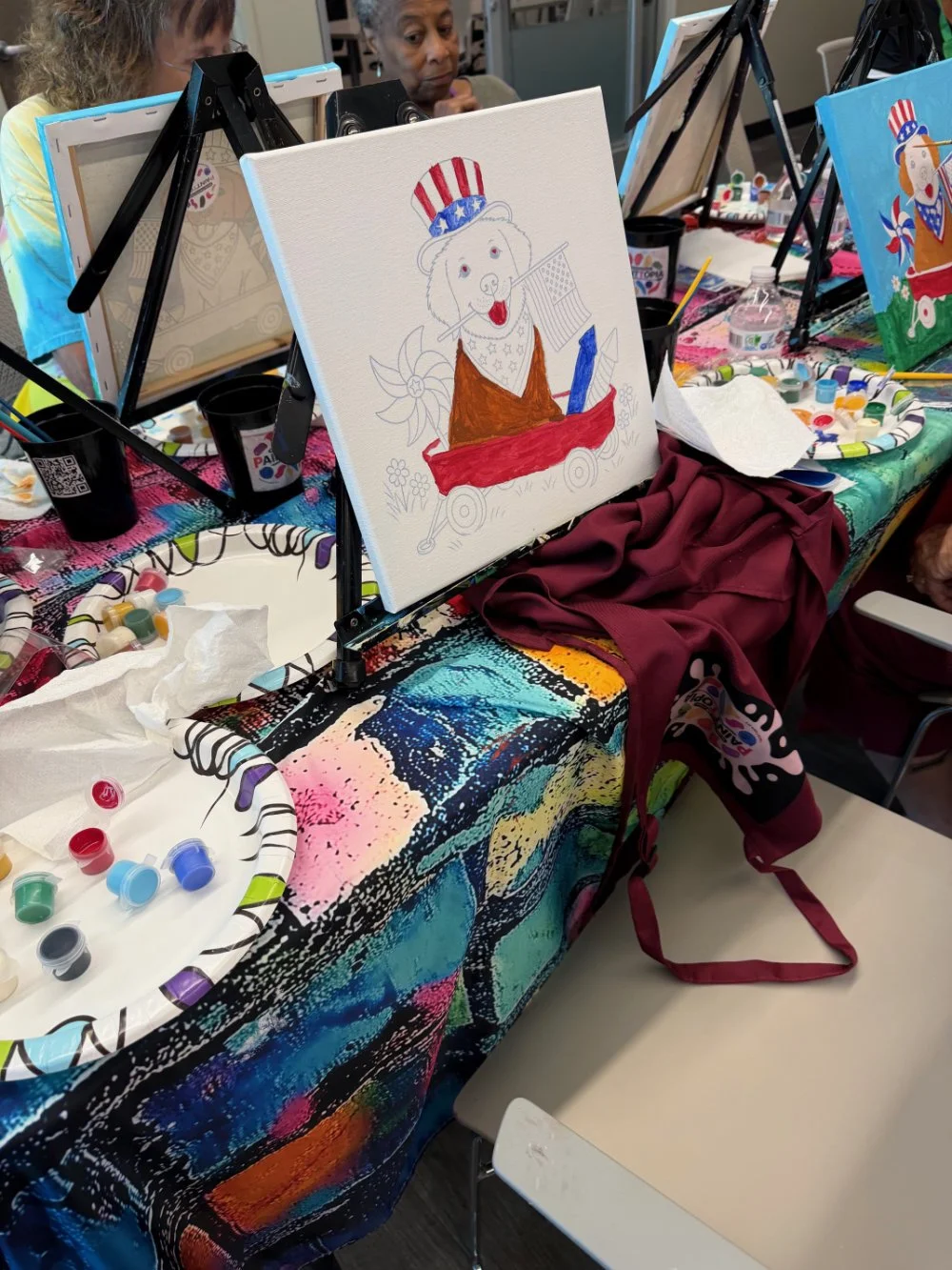 Canvas with a colorful drawing of a dog wearing an Uncle Sam hat, brown vest, and holding a flag, set up on an easel. The art supplies and paint bottles are scattered on a vibrant, multicolored tablecloth on the table.