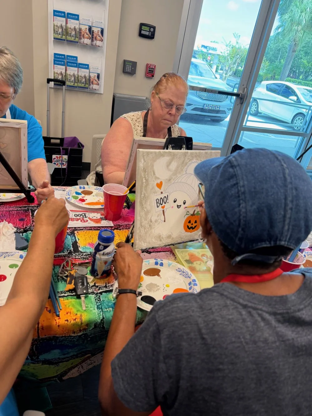 A group of people painting Halloween-themed canvases at a table covered with a colorful cloth. The scene is inside a room with large windows showing parked cars outside. One woman is wearing a blue apron, another woman has glasses and is focused on h