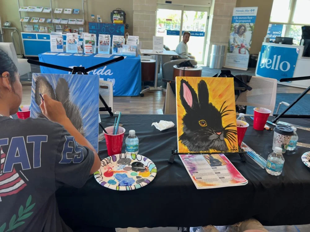 A person painting a black and gray cat on a canvas, with a painting of a black rabbit with pink ears on a stand next to them, at an indoor art event.