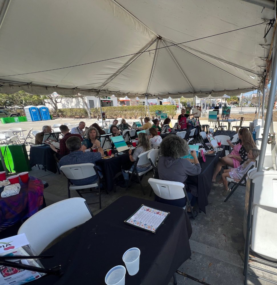 Group of people participating in a painting class under a large outdoor tent with painting supplies and easels.