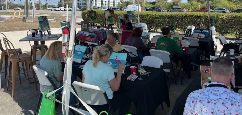 People painting tropical-themed pictures of flamingos and palm trees at an outdoor art event under a canopy on a sunny day.