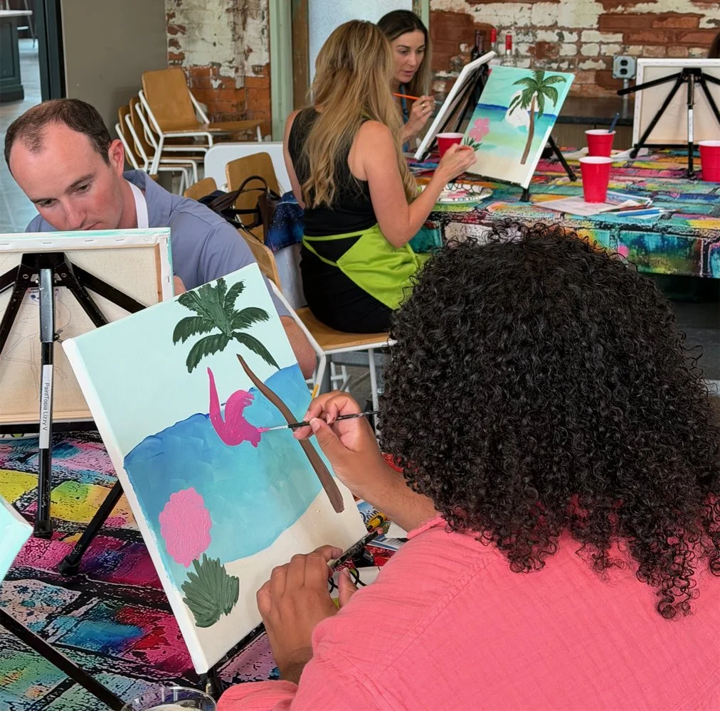 People participating in a paint and sip class, painting tropical beach scenes with palm trees, blue waters, and pink flamingos.