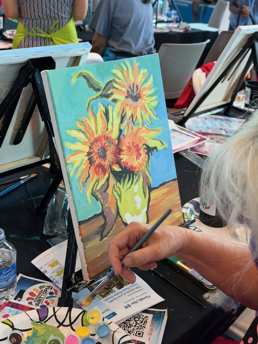 A person painting a colorful sunflower still life on a canvas at an outdoor art event.