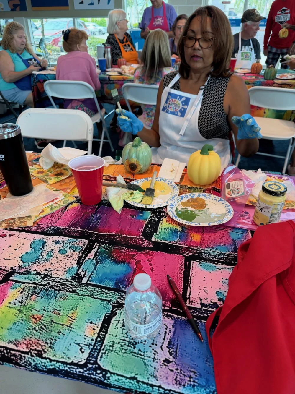 A woman wearing glasses, a black and white sleeveless top, an apron, and blue gloves is sitting at a colorful table with pumpkins, paint supplies, and a water bottle, participating in a painting activity. Behind her are several other women at a long 