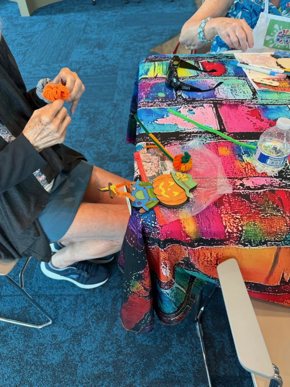 A person holding an orange craft item, sitting at a table with colorful splattered cloth, decorated with Halloween-themed craft supplies, including a pumpkin and a scarecrow cutout, with water bottle and sunglasses on the table.