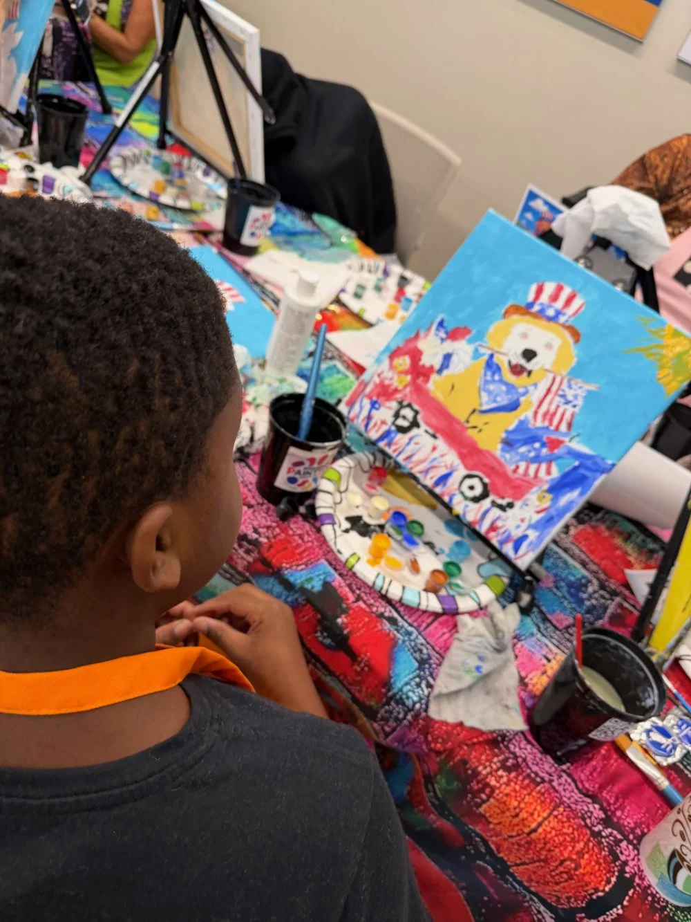 A child with short curly hair painting a colorful portrait of a dog wearing a Uncle Sam hat at an art station with paints, brushes, and supplies.