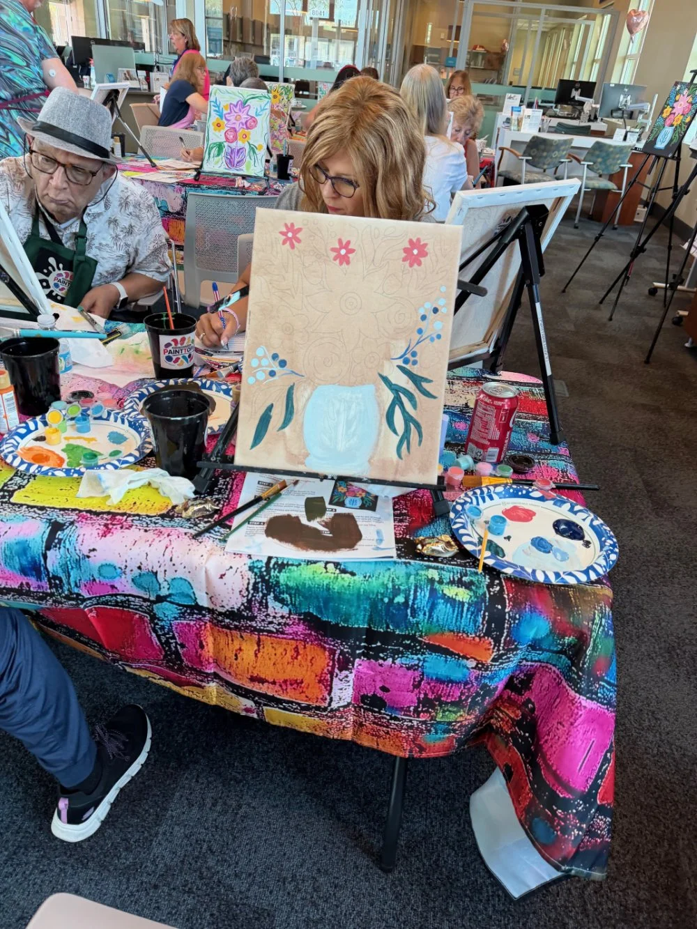 A group of people participating in a painting class at a colorful table with art supplies, paintings, and an unfinished floral artwork in progress.