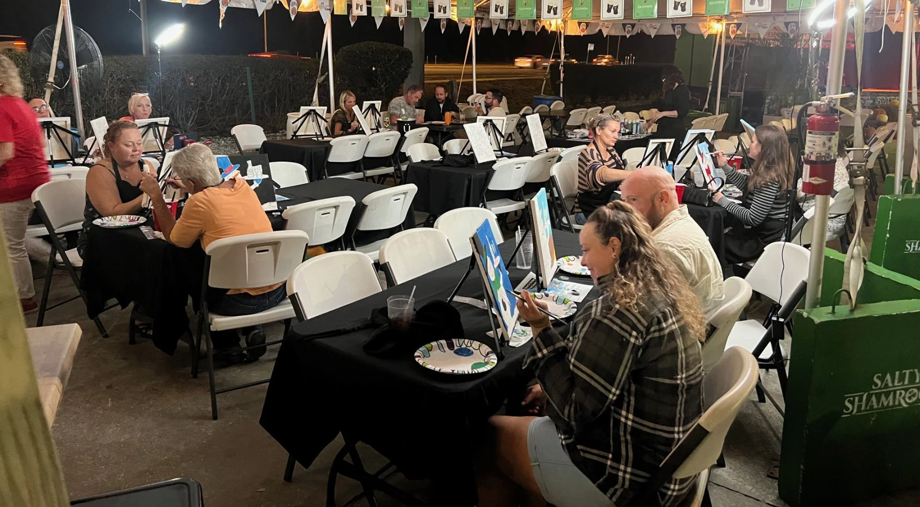 People painting and socializing at an outdoor painting event at night, with tables covered in black tablecloths and easels.