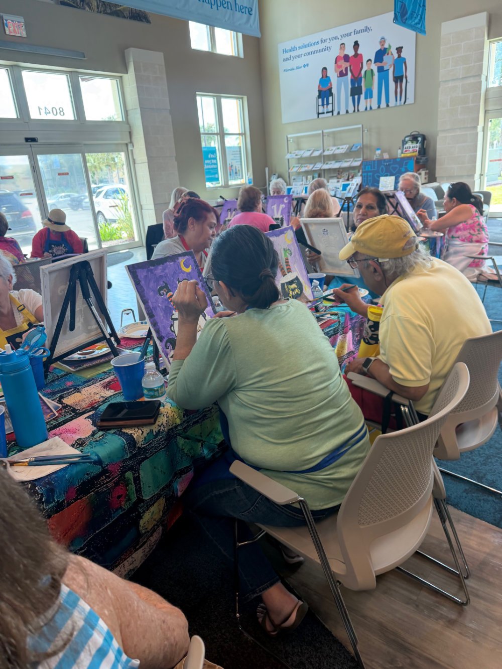 A group of people participating in a painting class indoors, seated at tables with painting supplies and easels, painting nighttime scenes with moon and stars.
