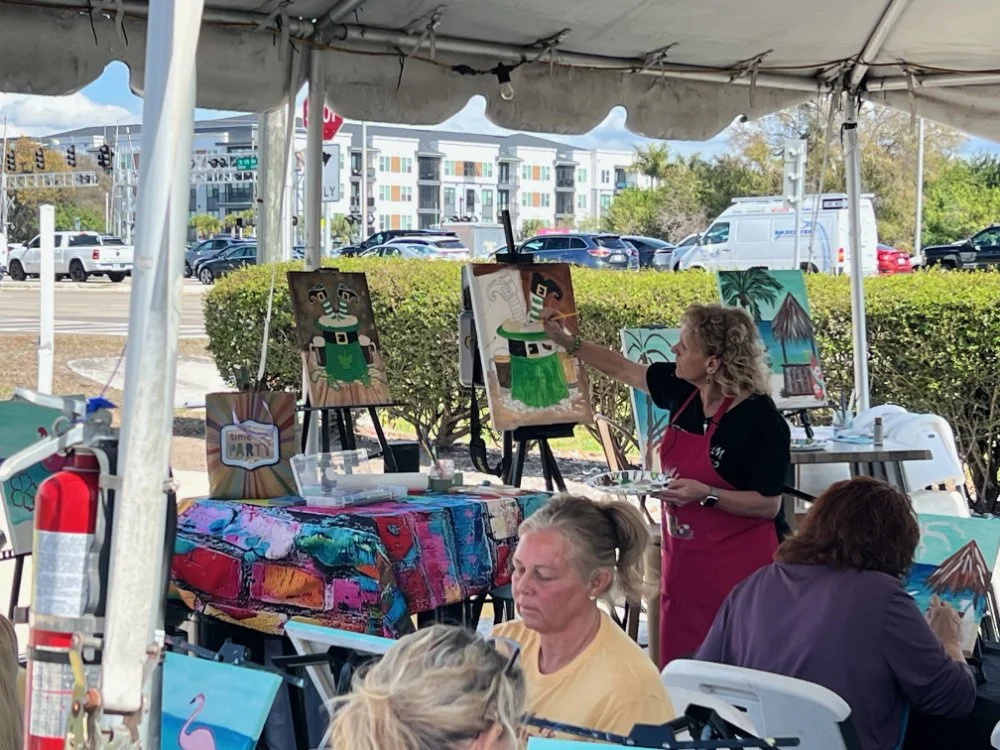 An outdoor art event where a woman is painting tropical-themed artworks, including a black and white figure in a green skirt, under a tent. Several people are seated, and a table with colorful cloth and art supplies is present. Background shows cars 