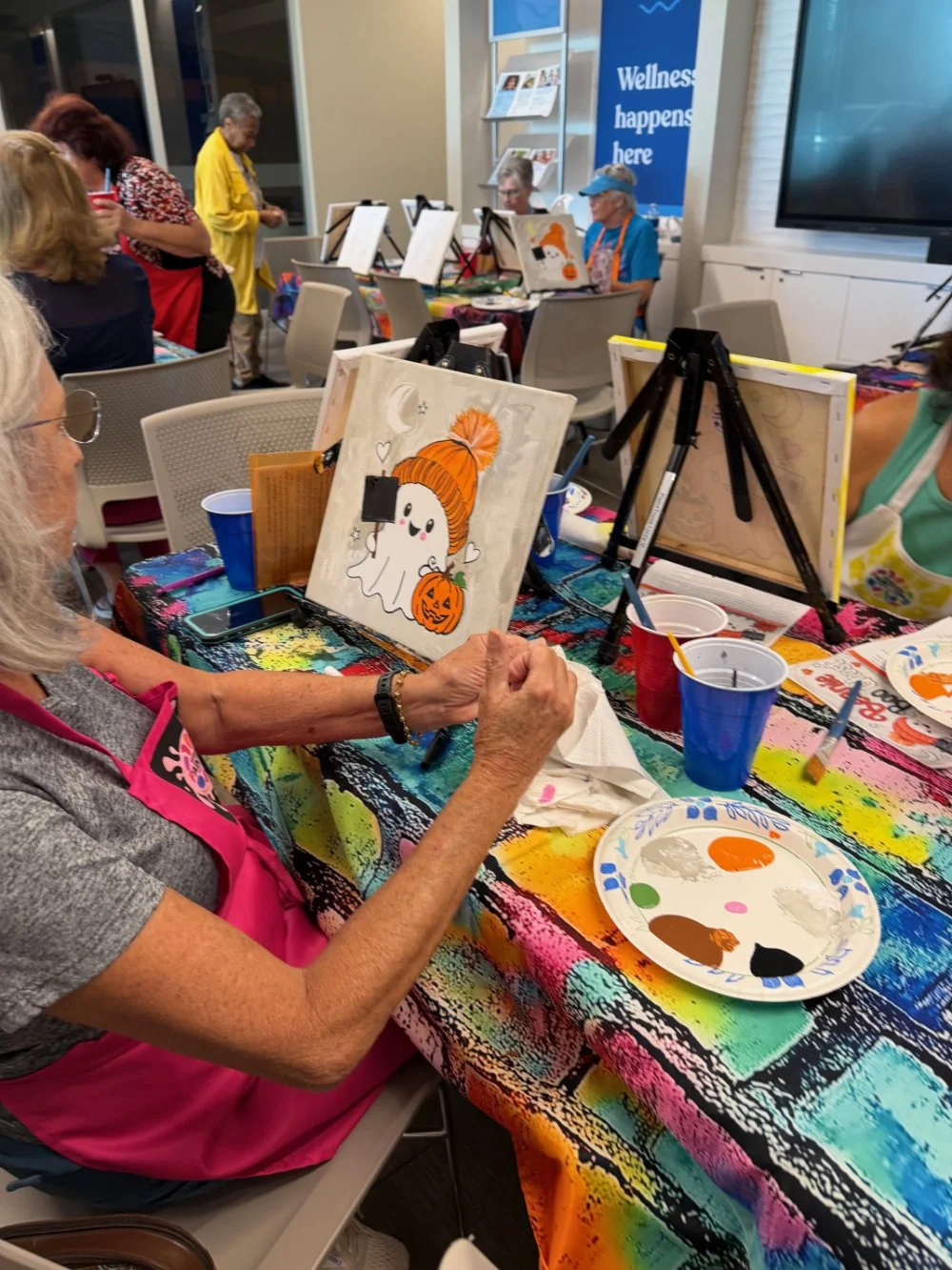 Older woman painting a Halloween-themed ghost with a pumpkin and orange hat, seated at a table covered with colorful paint splatters and painting supplies, at a group art event with other participants in the background.