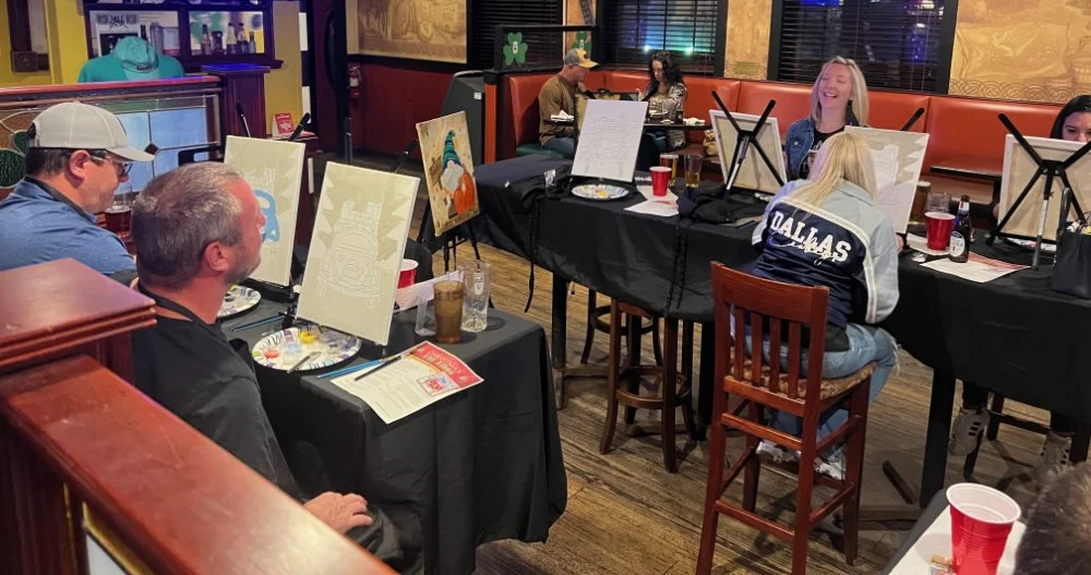 A group of people participating in a group painting event in a restaurant. They are sitting at tables with canvases, paint supplies, and drinks, smiling and engaged in painting.