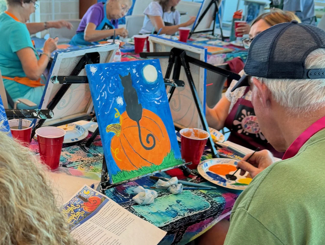 People participating in a group painting activity, creating a Halloween-themed picture of a black cat sitting on a pumpkin under a full moon with a night sky background, set up at tables with art supplies and drinks.